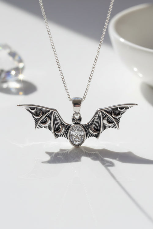 A silver bat pendant necklace with intricate wing detailing. hanging from a silver chain against a plain light gray background.