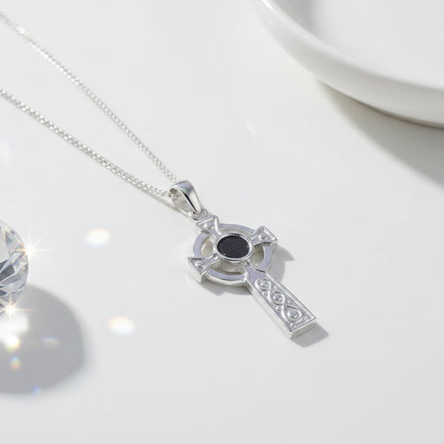 A sterling silver Celtic cross pendant necklace with a black center stone, resting on draped cream-colored satin fabric.